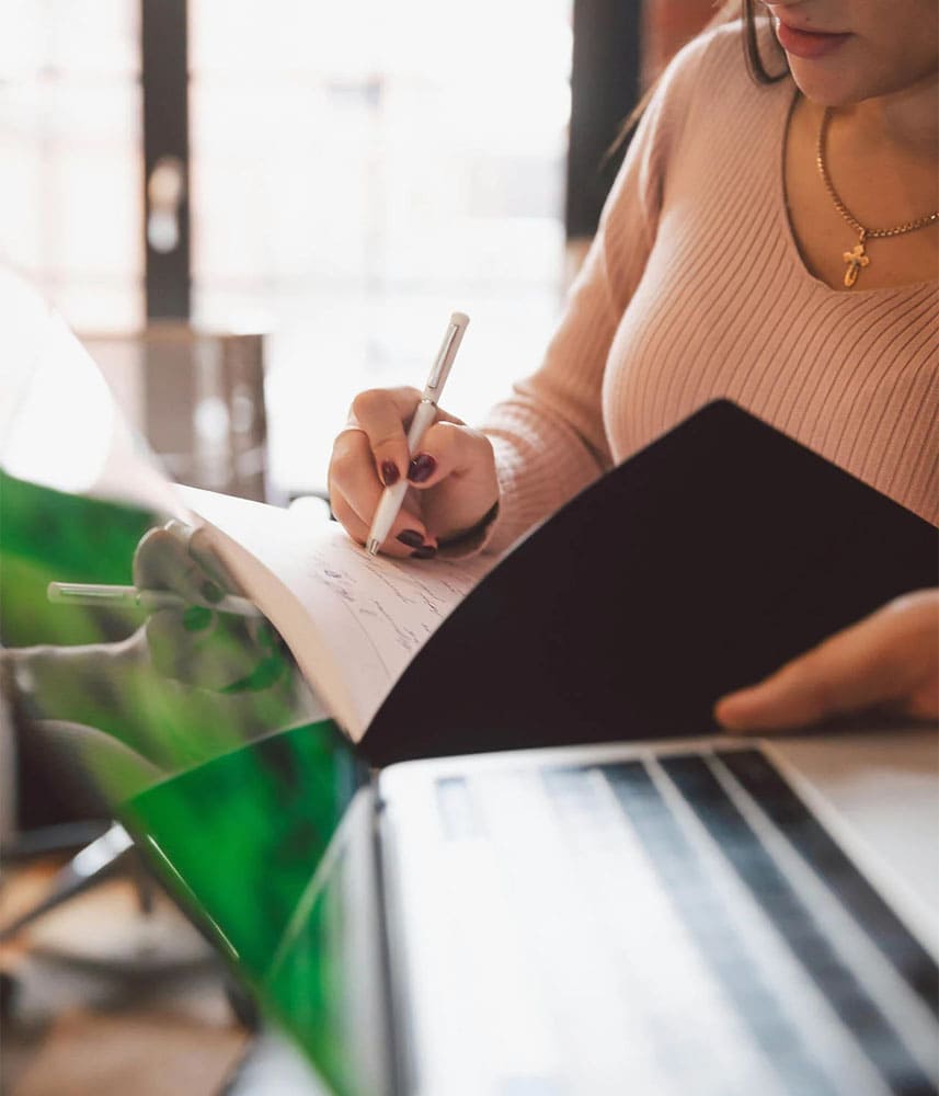 Femme écrivant dans un cahier, devant un ordinateur portable